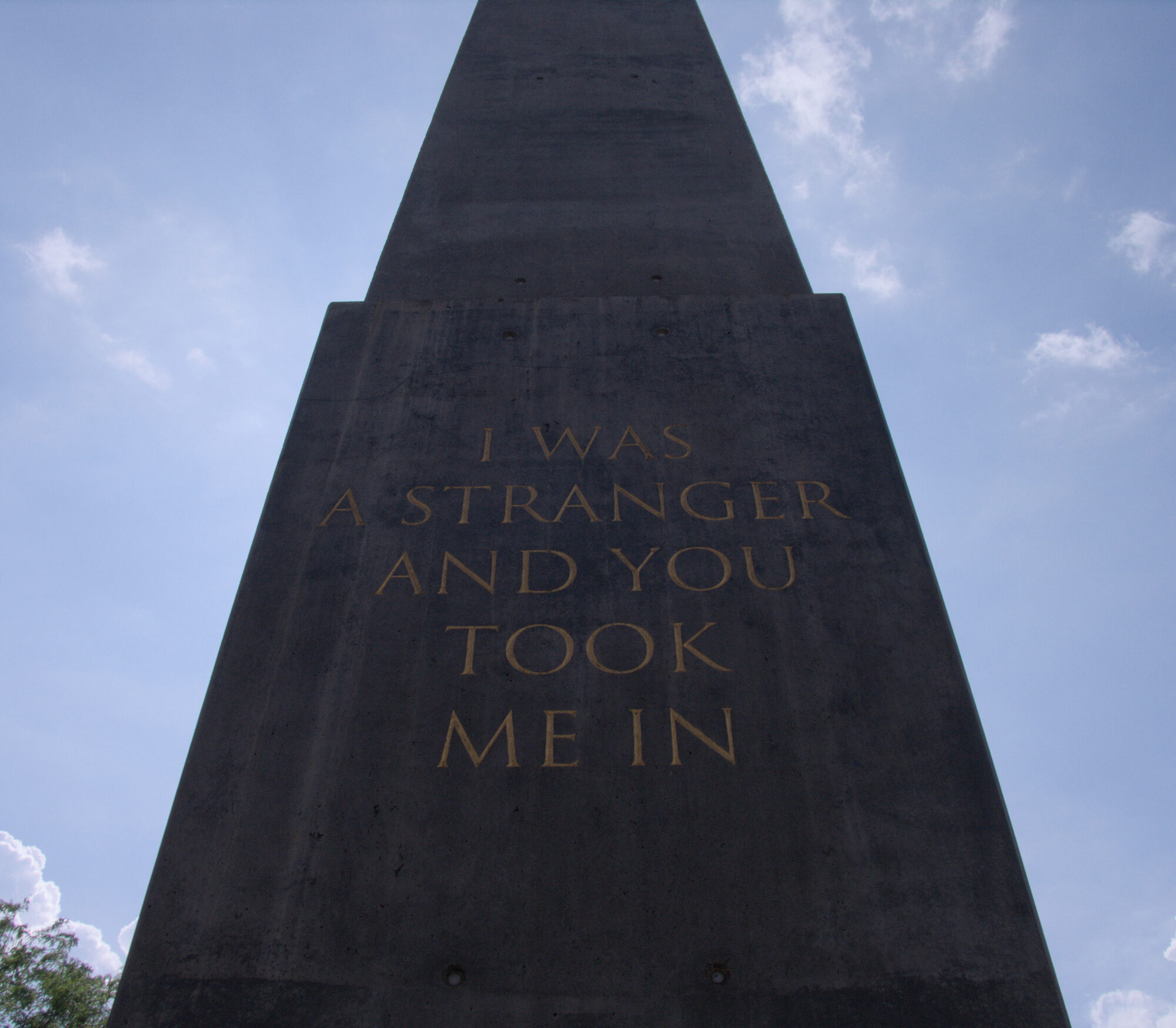 An obelisk of dark concrete seen from below, incised in gold capitals: I WAS A STRANGER AND YOU TOOK ME IN